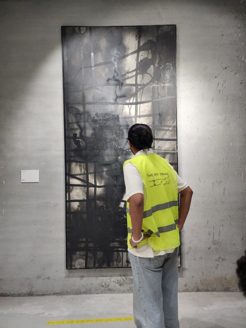 A person (Awais Shaukat) wearing a bright yellow reflective vest printed with “THE NCA TRIENNALE” stands with hands on hips, closely observing a tall, dark abstract painting mounted on a bare concrete wall in a gallery space.